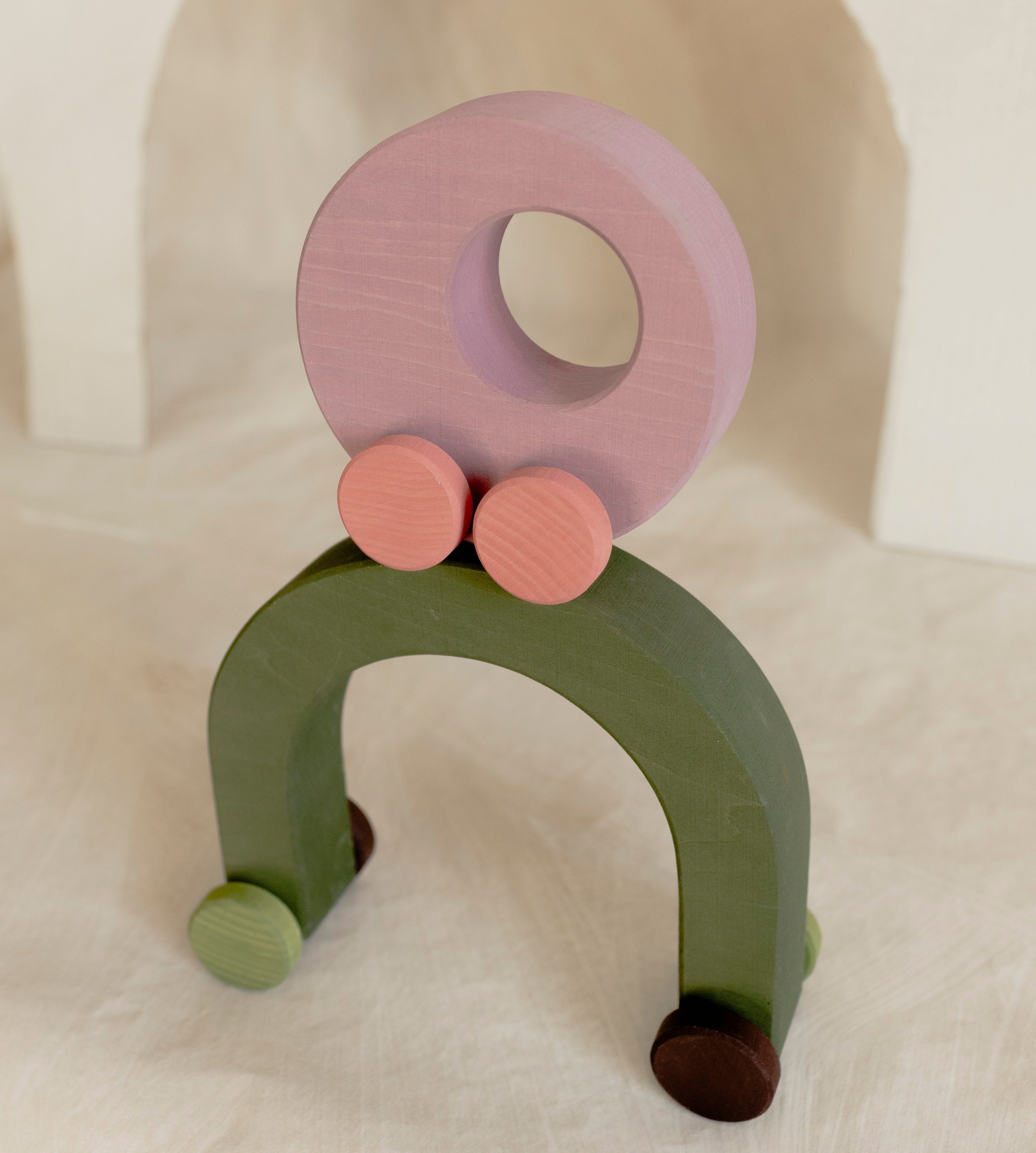 A minimalist wooden toy arch with a pastel green base and a lavender-purple circular top, accented with small pink and brown wheels, set against a warm beige background.